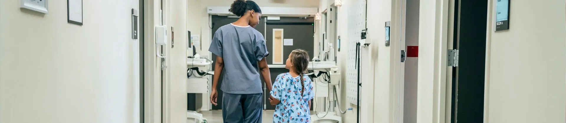 woman nurse walking down hospital hallway holding hand of girl in hospital gown