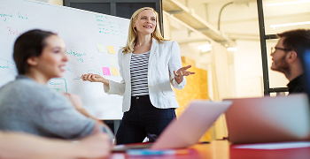 woman in a white blazer talks to to colleagues in front of white board