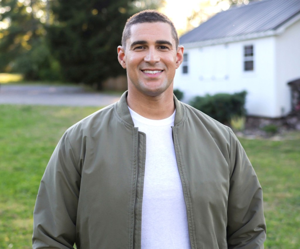 guy in white shirt and grey jacket standing in grass in front of home